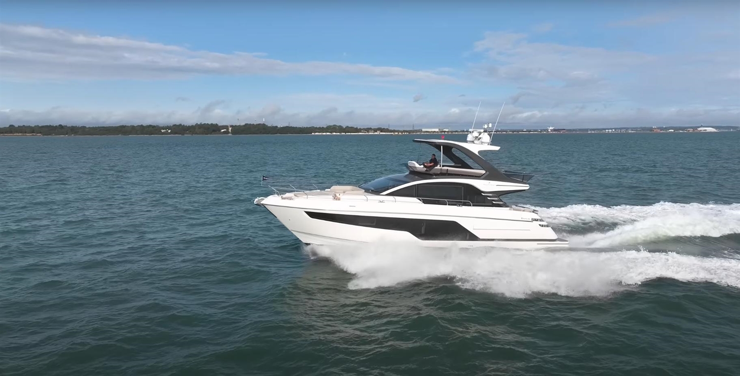 Luxury motor yacht cruising on blue water, creating a white wake, with a clear sky and shoreline in the background.
